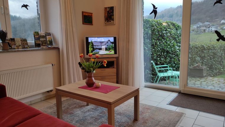 A cozy living room with a red sofa and a wooden table. Through the windows, one can see a green terrace and the surrounding landscape.