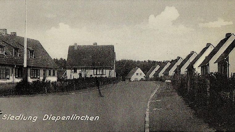 Eine ruhige Straße mit Wohnhäusern in einer Siedlung. Die Umgebung ist grün und ländlich.