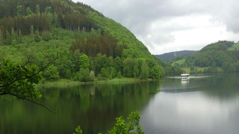 A peaceful lake surrounded by green hills and trees. In the background, a boat can be seen.