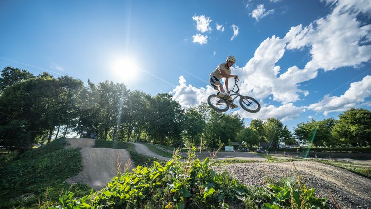 A BMX rider jumps over a ramp in a park. The sun is shining brightly in the sky, surrounded by trees and clouds.