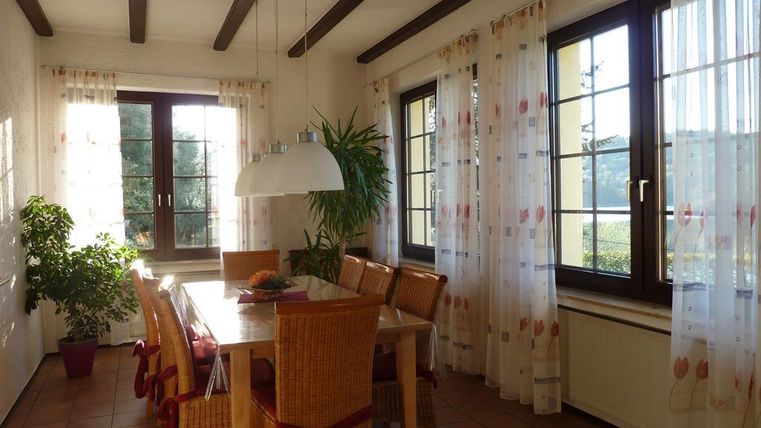 A bright dining room with a large table and chairs. The windows are decorated with curtains, and a plant is standing in the corner.