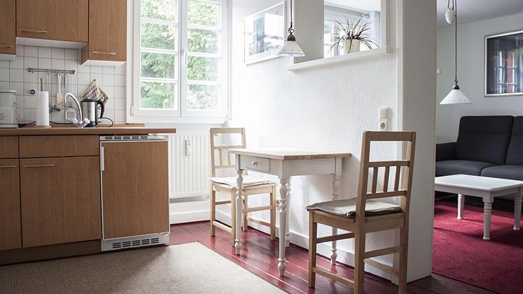 Een gezellige keuken met houten kasten en een kleine eetruimte. Een lege tafel met twee stoelen en een zitgedeelte op de achtergrond.