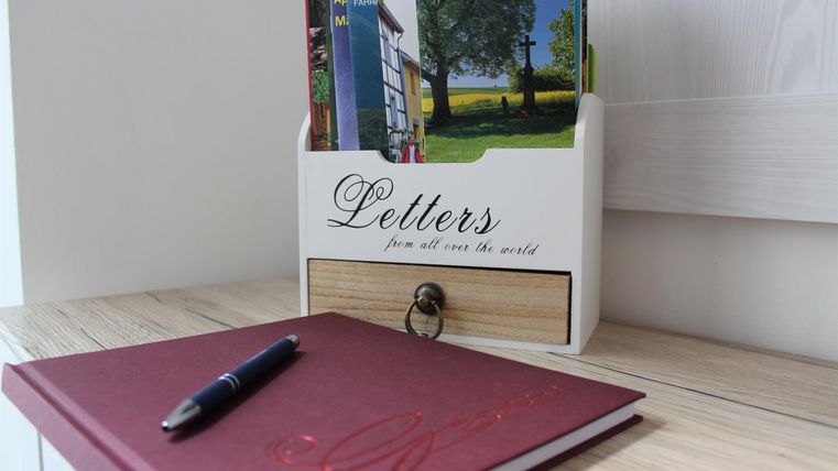 A wooden shelf with postcards and a notebook on a table. Next to it, there is a ballpoint pen.