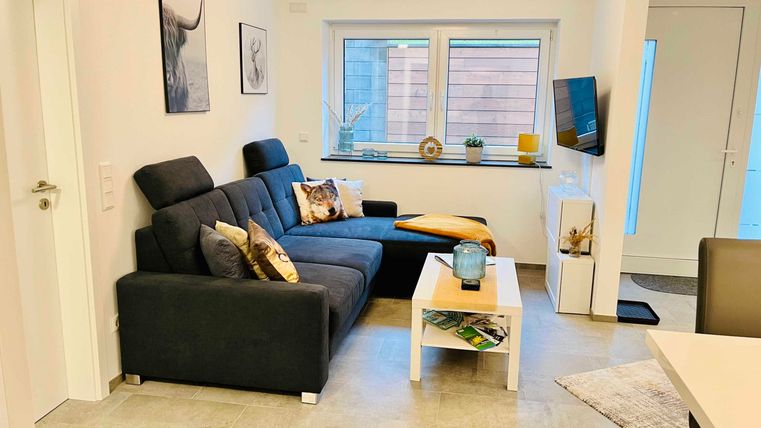 A modern living room with a dark gray couch and a small table. Bright walls and large windows create a friendly atmosphere.