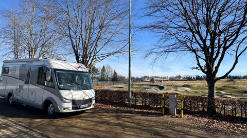 Een camper staat op een parkeerplaats tussen bomen. Op de achtergrond is een uitgestrekt weiland te zien.