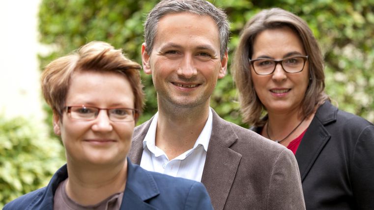 Three people are smiling in front of a green backdrop. They are wearing formal attire and appear friendly and professional.