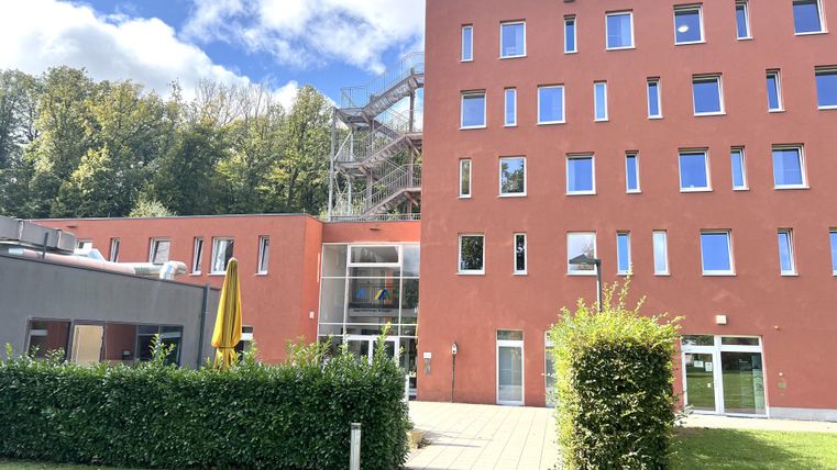 Ein modernes, orangefarbenes Gebäude steht in einer grünen Umgebung. Im Hintergrund sind Bäume und ein blauer Himmel sichtbar.