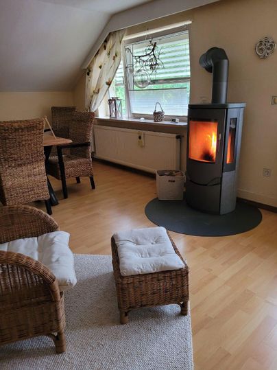 Ein gemütliches Wohnzimmer mit einem Holzofen und Rattanstühlen. Das Fenster lässt viel Tageslicht herein und sorgt für eine einladende Atmosphäre.