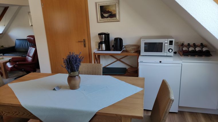 A cozy dining area with a table, chairs, and a vase full of lavender. In the background, there is a microwave, a coffee machine, and shelves with wine bottles.