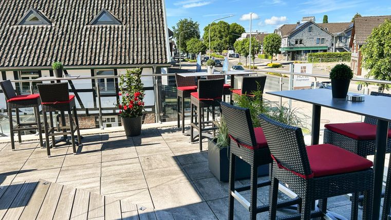 A modern terrace with tables and chairs in a restaurant. In the background, there are houses and trees under a clear blue sky.