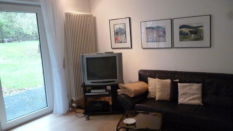 Een gezellige woonkamer met een zwarte bank en een televisie. Grote ramen laten veel daglicht binnen en bieden uitzicht op de tuin.