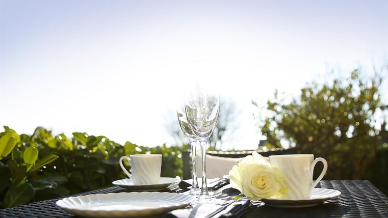 An elegantly laid table outdoors with white cups, glasses, and a bouquet of flowers. The blue sky and green foliage create a fresh atmosphere.