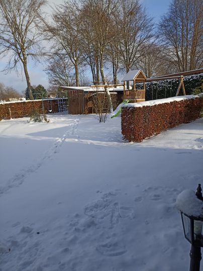 Ein Garten im Schnee mit einem Spielplatz und einigen Bäumen. Die Landschaft ist ruhig und winterlich, mit Fußspuren im Schnee.