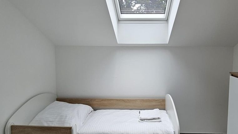 Een eenvoudige slaapkamer met een eenpersoonsbed en een klein raam. De muren zijn wit en de vloer is grijs.