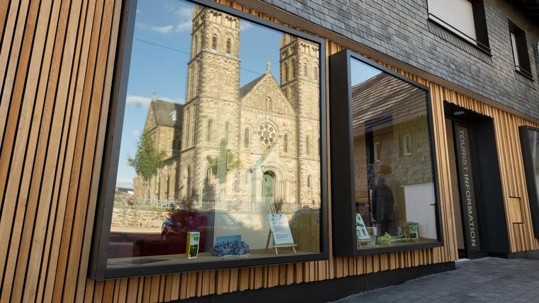 A modern building with wooden elements and large windows reflects a beautiful church. The church has two towers and is surrounded by trees.