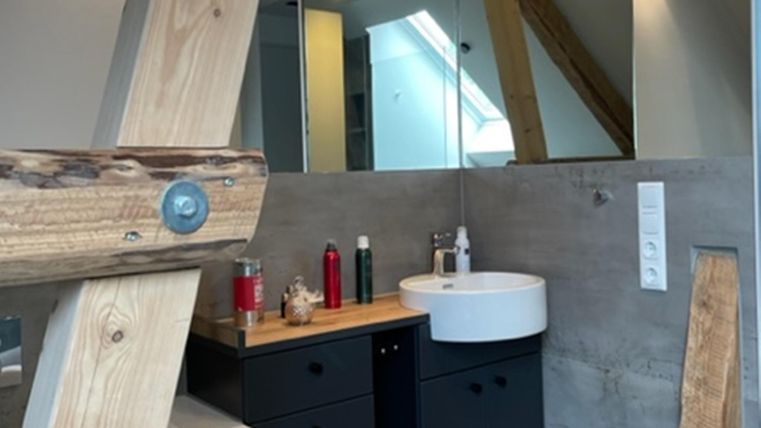 Een moderne badkamer met een ronde wastafel en houten plank. De muren zijn van beton, en er zijn grote ramen die veel licht binnenlaten.
