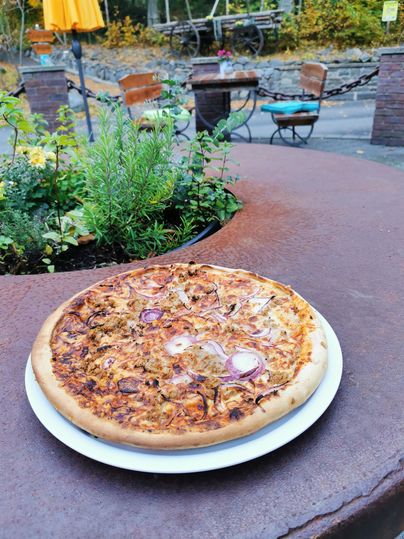 Een verse pizza op een rustieke tafel. Op de achtergrond zijn enkele zitplaatsen en planten zichtbaar.