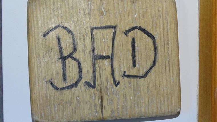 Een houten bord met de tekst "BAD". Het hangt aan een muur en geeft de toegang tot een badkamer aan.