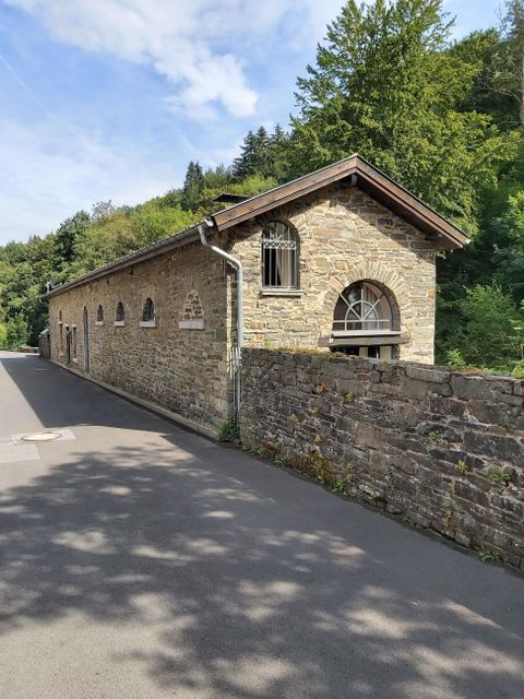 A historic stone building with a gabled roof, surrounded by trees. The street runs directly past the building.