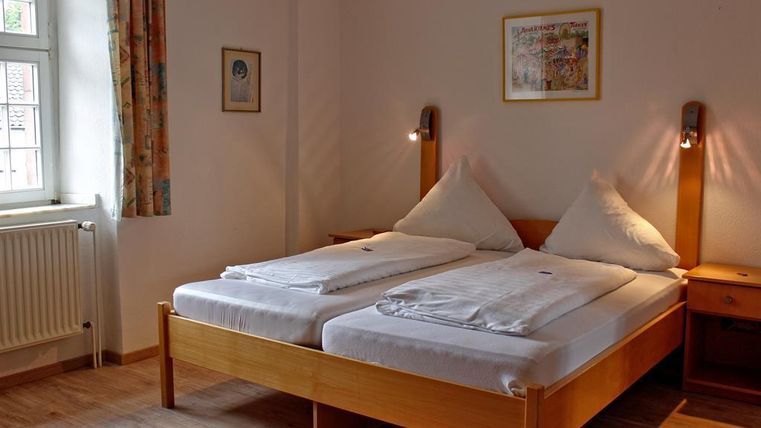 Een lichte kamer met twee eenpersoonsbedden en houten meubels. De muren zijn neutraal geschilderd en de kamer heeft grote ramen die voor natuurlijk licht zorgen.