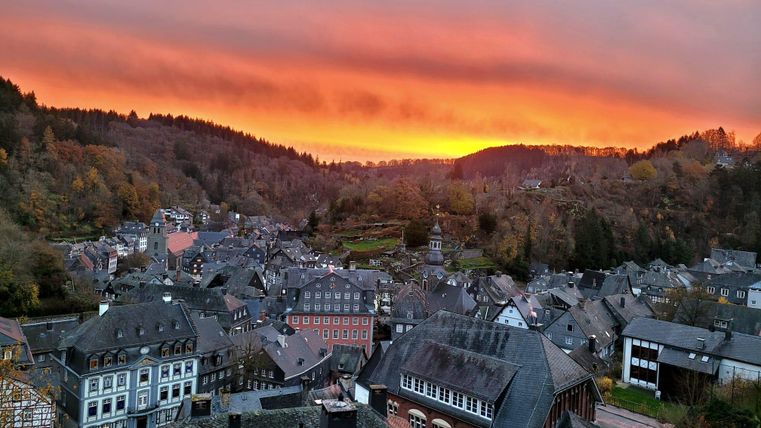 Ein malerisches Dorf mit alten Gebäuden, umgeben von bewaldeten Hügeln. Der Sonnenuntergang färbt den Himmel orange und pink.