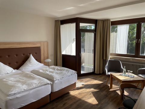 Een heldere hotelkamer met twee eenpersoonsbedden, een tafel en stoelen. Grote ramen zorgen voor veel daglicht.