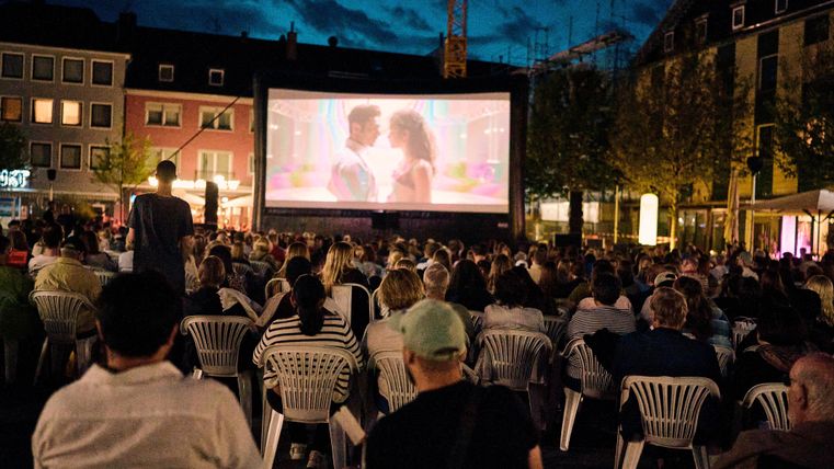 Ein Freiluftkino mit einer großen Leinwand, auf der ein Film gezeigt wird. Viele Menschen sitzen auf weißen Stühlen und genießen die Vorstellung bei Nacht.