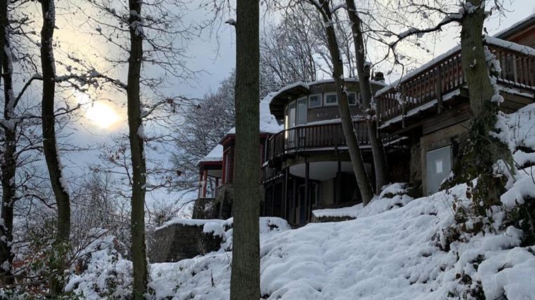A snowy landscape with a house standing on a hill. The sun is shining weakly through the clouds.