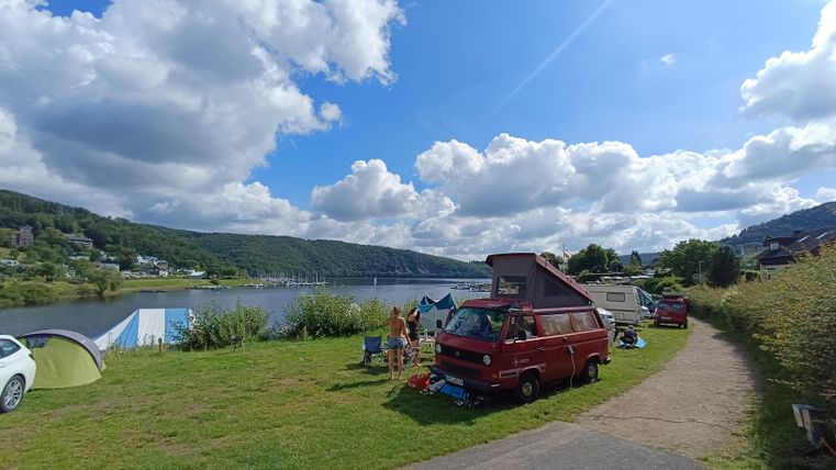 A beautiful campsite by the river with a red RV. The landscape is surrounded by green meadows and a blue sky with fluffy clouds.