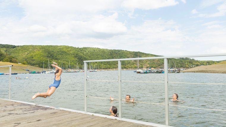 Een kind springt van een steiger in het water, terwijl andere mensen aan het zwemmen zijn. Op de achtergrond zijn groene heuvels en een heldere lucht te zien.