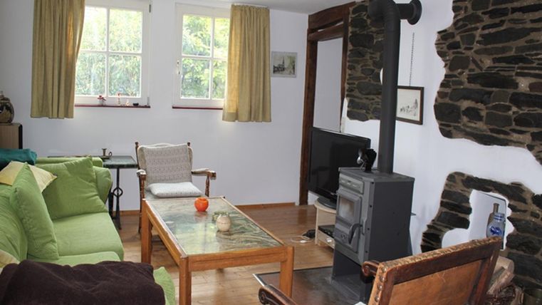 A cozy living room with a wood stove and a large window front. The decor includes comfortable sofas and a wooden table.
