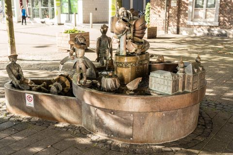 A creative fountain installation with bronze-like figures and various motifs. The scene depicts playing children and a romantic cityscape.