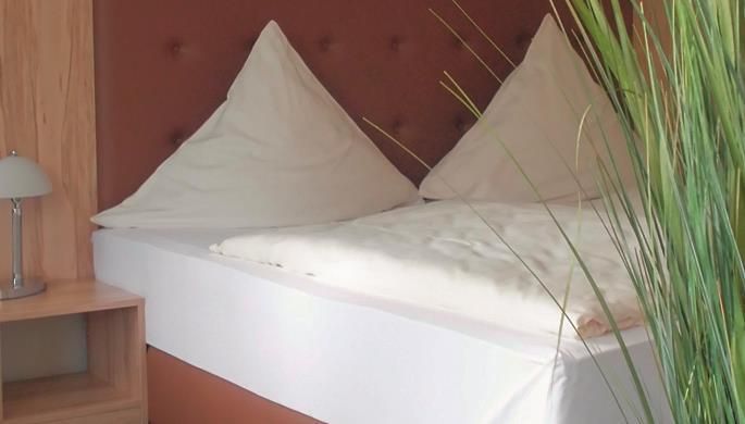 Een gezellig tweepersoonsbed met witte kussens en een lichte dekbed. Naast het bed staat een nachtkastje met een lamp en groene planten op de achtergrond.