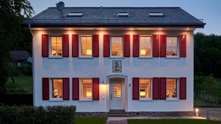 Een modern, twee verdieping tellend huis met witte muren en rode shutters. De ramen zijn verlicht en er is een smal pad naar de voordeur.