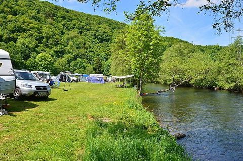 Ein grünes Campinggebiet neben einem ruhigen Fluss. Es gibt Wohnmobile und Zelte, umgeben von Bäumen und Wiese.