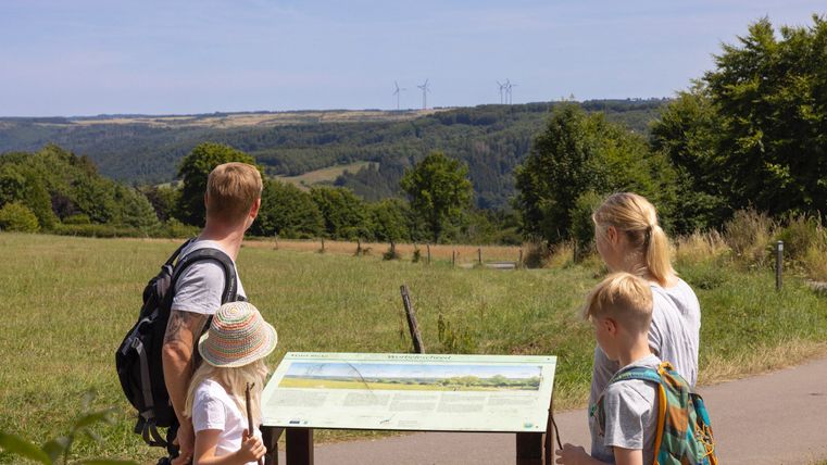 A family stands in front of an informational sign in a green landscape. In the background, rolling fields and trees can be seen under a clear sky.
