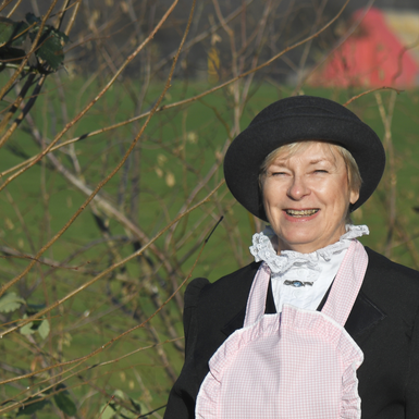 Eine lächelnde Frau trägt einen Hut und eine Schürze. Im Hintergrund sind grüne Bäume und Sträucher zu sehen.