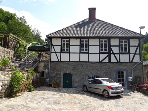Een historisch huis in vakwerkstijl met een geplaveide binnenplaats. Op de voorgrond staat een grijze auto, terwijl de tuin op de achtergrond groen is.