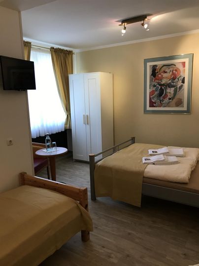 Een eenvoudige kamer met twee bedden, een televisie en een kast. De kamer heeft grote ramen met gordijnen en is licht ingericht.