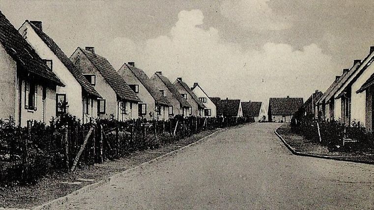 Eine ruhige Straße mit kleinen, weißen Häusern auf beiden Seiten. Der Himmel ist bewölkt und die Umgebung wirkt ländlich.