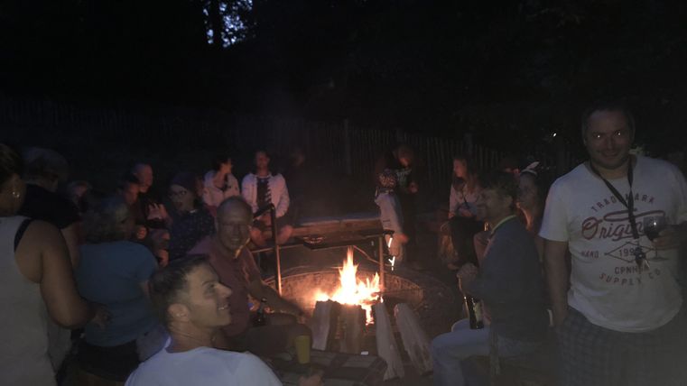 Een groep mensen zit om een kampvuur in een donkere omgeving. Er heerst een gezellige en sociale sfeer.