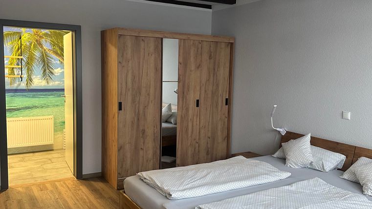 A modern bedroom with a large double bed and a wooden wardrobe. In the background, there is a window overlooking a sunny landscape.