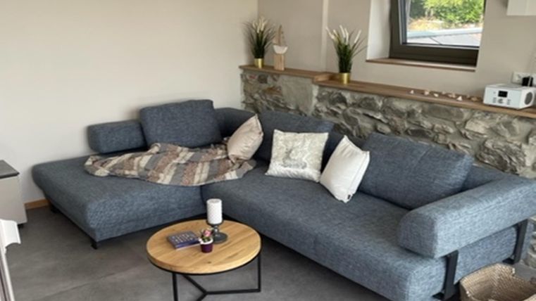 Een modern woonkamer met een grijze bank en een ronde houten tafel. Op de achtergrond bevindt zich een stenen muur en een grote raampartij.