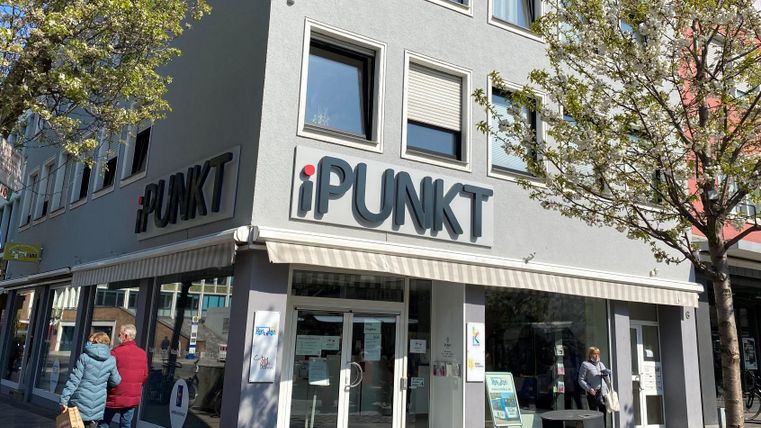 A modern store named "iPUNKT" in an urban setting. People walk by and there are trees providing shade.