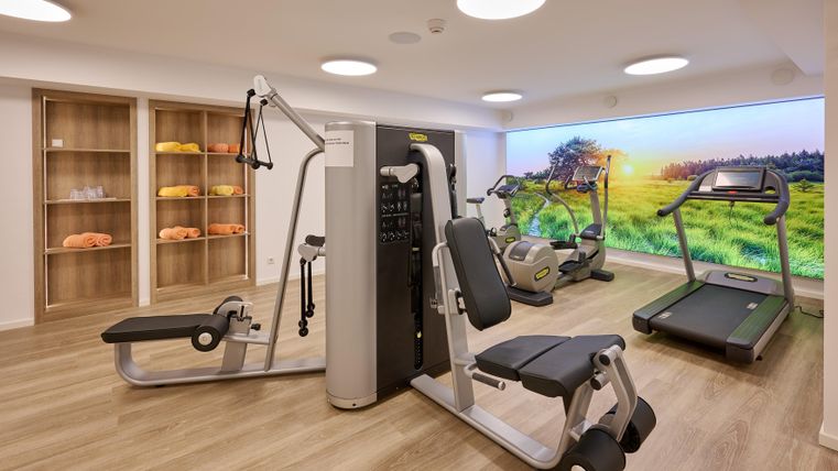 Ein modernes Fitnessstudio mit verschiedenen Geräten. Im Hintergrund ist eine große Wand mit einem Bild einer grünen Landschaft zu sehen.