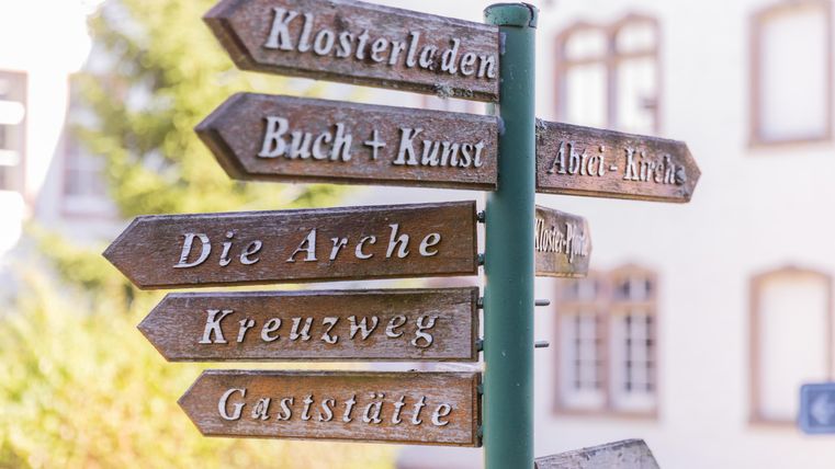 Ein Wegweiser mit verschiedenen Richtungen. Die Schilder zeigen Orte wie „Klosterladen“ und „Buch + Kunst“.