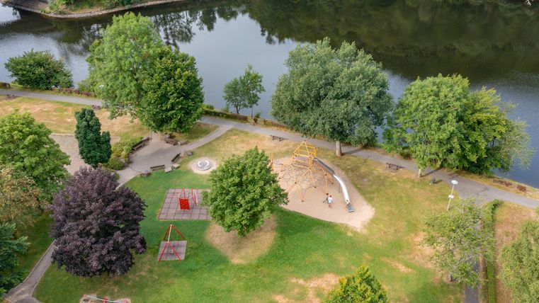A green park by the water with many trees. In the park, there are playground equipment and seating options.