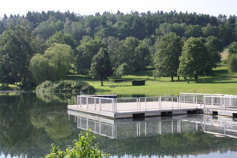 Een rustige meer met een houten platform op de voorgrond. Op de achtergrond zijn groene bomen en zachte heuvels te zien.
