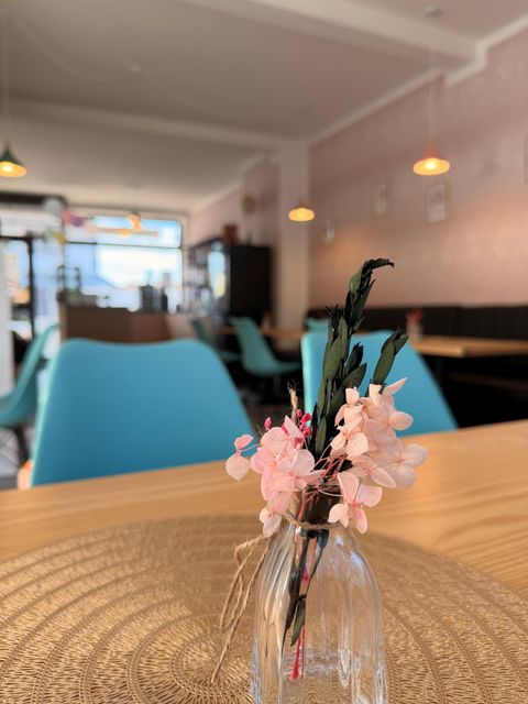 A cozy café with blue chairs and warm lighting. A small glass vase with pink flowers is on the table.