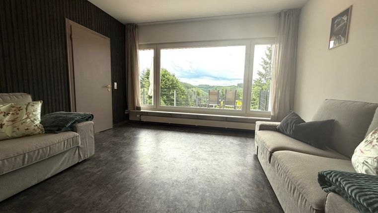 Een moderne woonkamer met grote ramen en uitzicht op de natuur. De meubels zijn eenvoudig en stijlvol ontworpen.